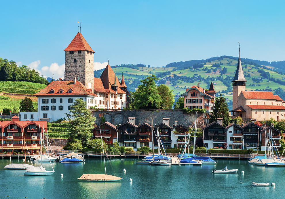 Spiez Castle and church overlooking a marina filled with sailboats on the turquoise waters of Lake Thun, with vineyards and green Swiss Alpine hills in the background.