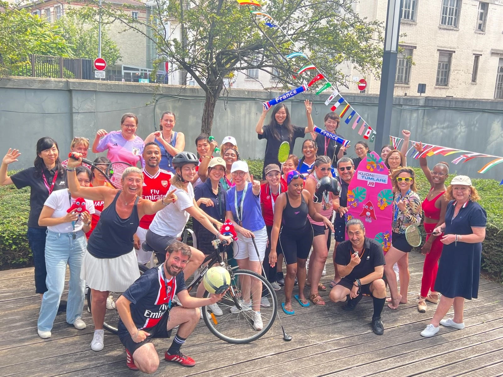 파리 2024 올림픽 기간, 다양한 국적의 게스트들과 쿠오니 투믈라레 DMC 팀이 국기 스카프와 테니스 라켓, 브랜드 사인을 들고 야외에서 밝게 단체 사진을 찍고 있다.