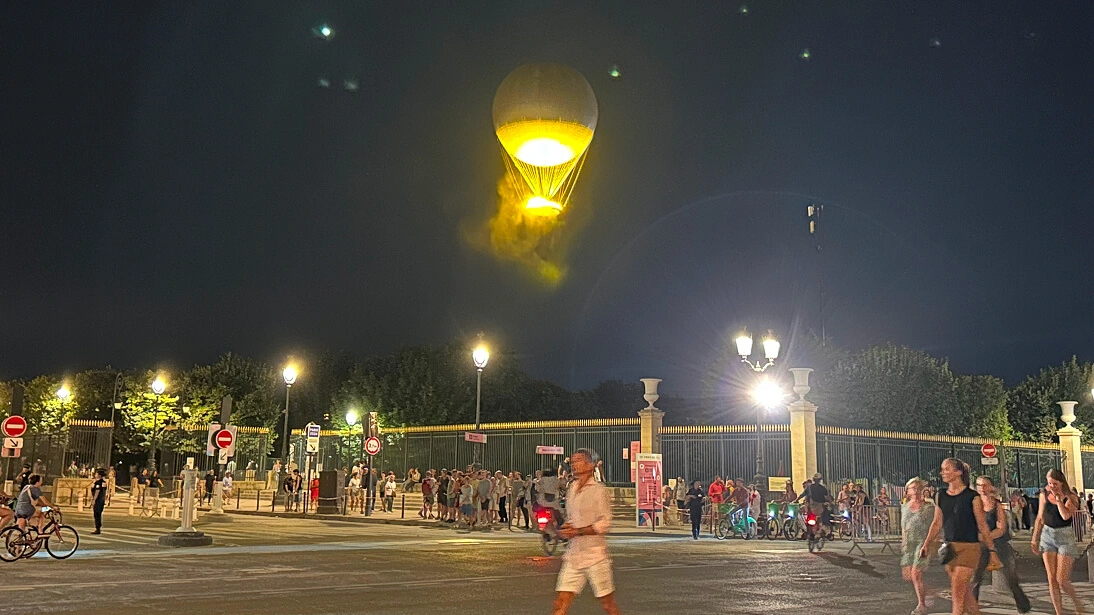파리 야간 풍경으로, 거대한 조명된 올림픽 성화대 풍선이 튈르리 정원 위에 떠 있고, 많은 사람들이 거리에 모여 있으며, 가로등이 주변을 밝히고 있다.