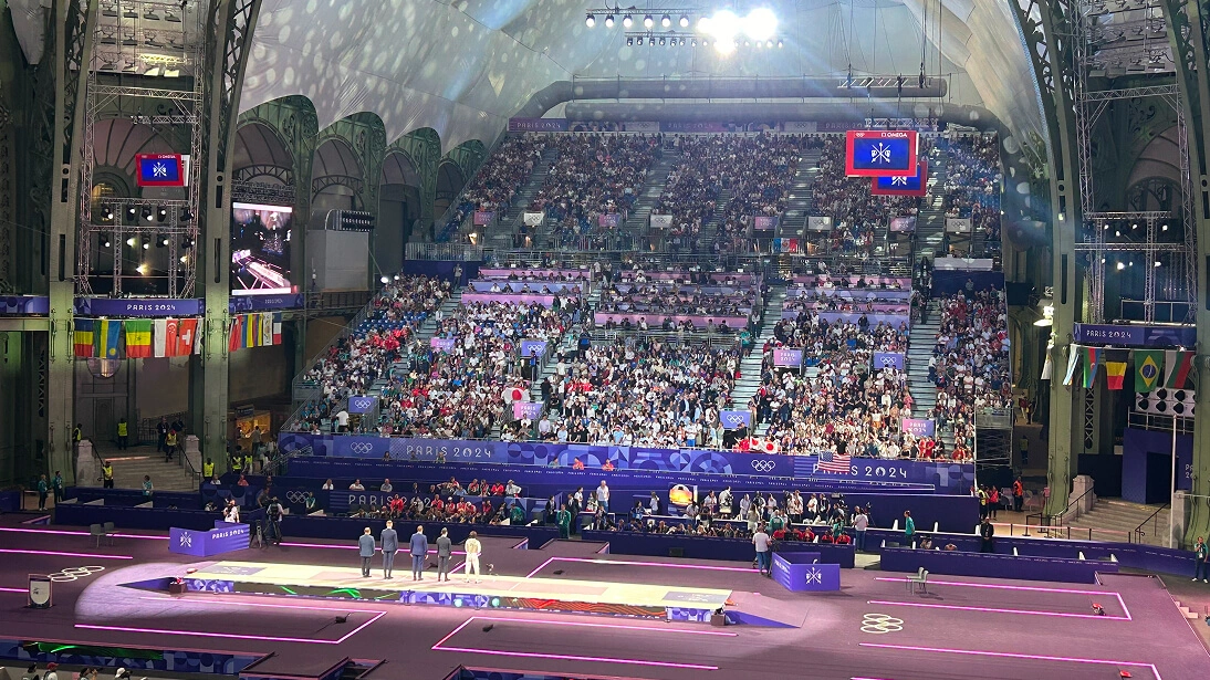 파리 2024 올림픽 기간, 그랑 팔레 경기장 만석. 체조 선수들이 경기장에서 경기 중이며, 각국 국기와 관중으로 가득한 관람석.