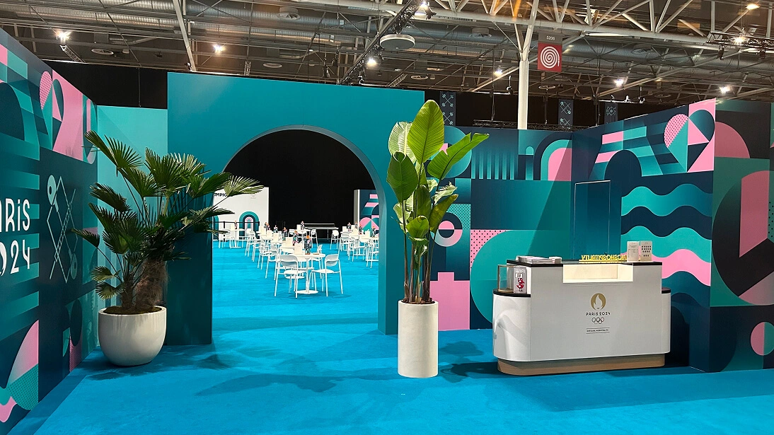 화려한 파리 2024 올림픽 브랜딩 입구. 청록색 아치, 화분, 파란 카펫 위 리셉션 데스크.