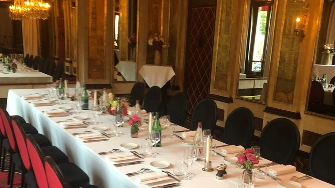 화려한 금색 벽과 거울이 있는 고급 다이닝룸. 긴 테이블에 흰 린넨, 정갈한 세팅, 플라워 센터피스와 검은 의자가 놓여 있다.