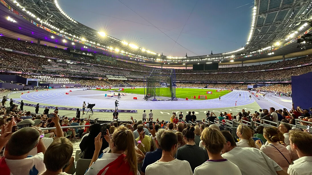 파리 2024 올림픽 해질녘의 스타드 드 프랑스. 밝은 조명 아래 만석 관중이 환호하며 보라색 트랙과 필드를 내려다보고 있다.