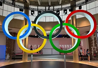 조명이 밝혀진 올림픽 오륜기가 실내에 걸려 있고, 관람객들이 아래를 지나가고 있다.