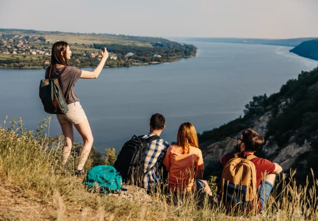 Student travellers with backpacks on a hilltop taking photos of a sweeping river-valley view, gaining new perspectives.