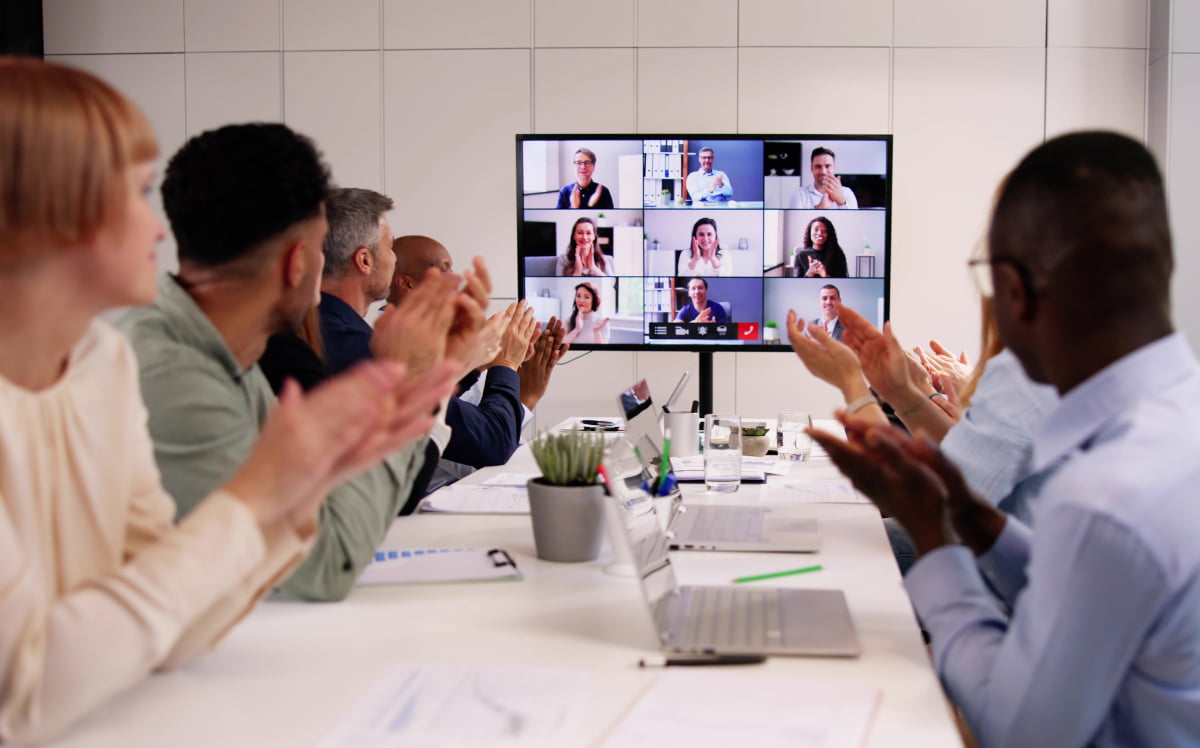 Team members in a meeting room applaud colleagues joining by video call on a large wall-mounted screen.