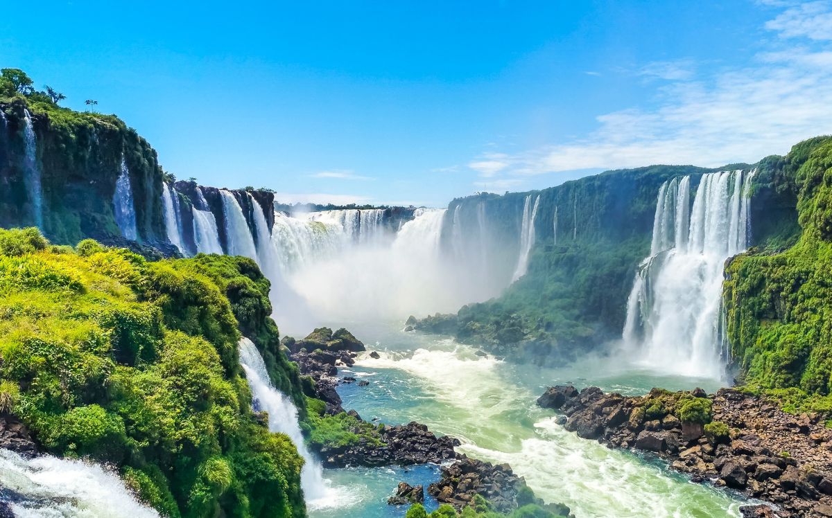 Panoramic view of the Iguazu Falls cascading through tropical rainforest on the Brazil–Argentina border, under a blue sky.