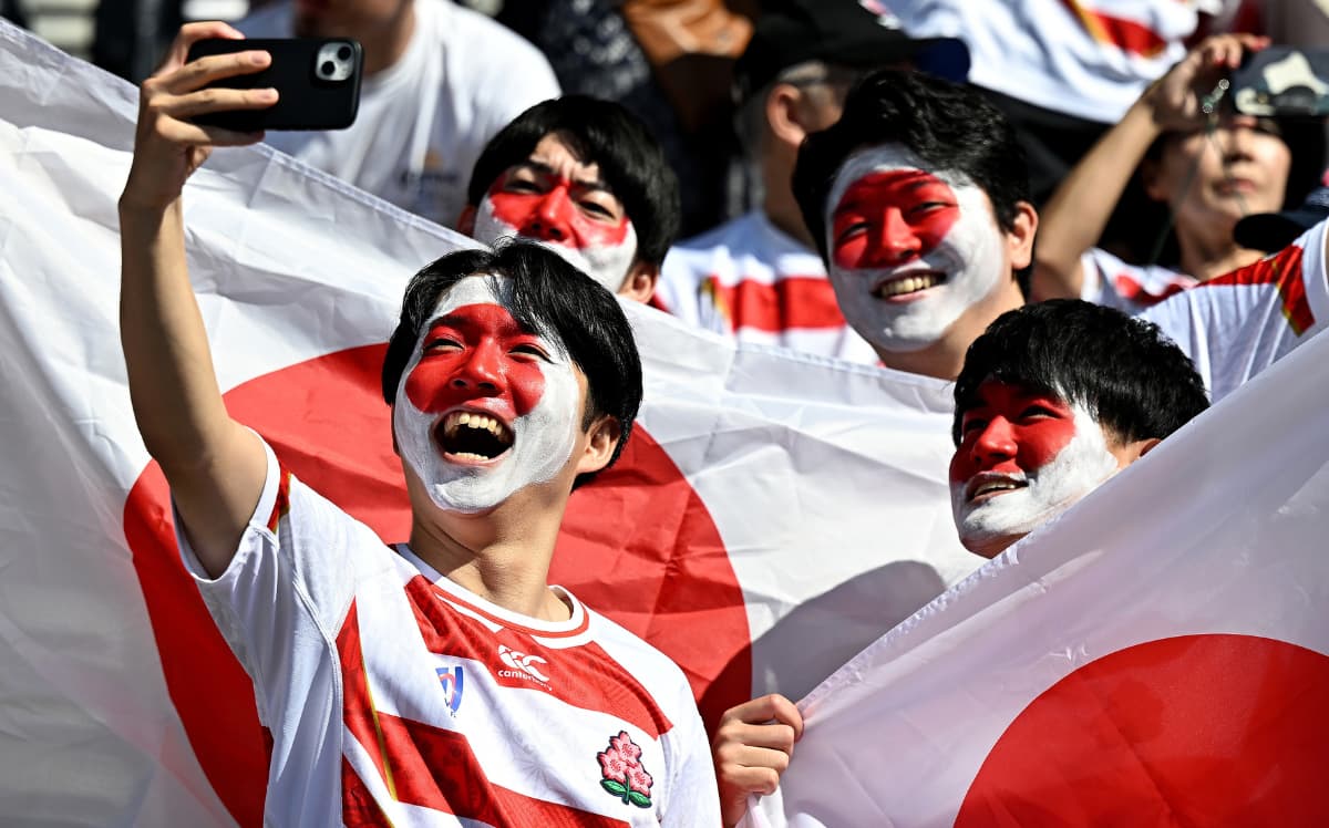 Japan rugby fans with Japanese flag face paint cheer and take a selfie in front of large Japanese flags in a stadium.