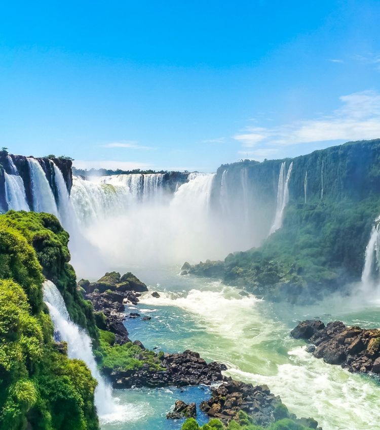 Panoramic view of the Iguazu Falls cascading through tropical rainforest on the Brazil–Argentina border, under a blue sky.