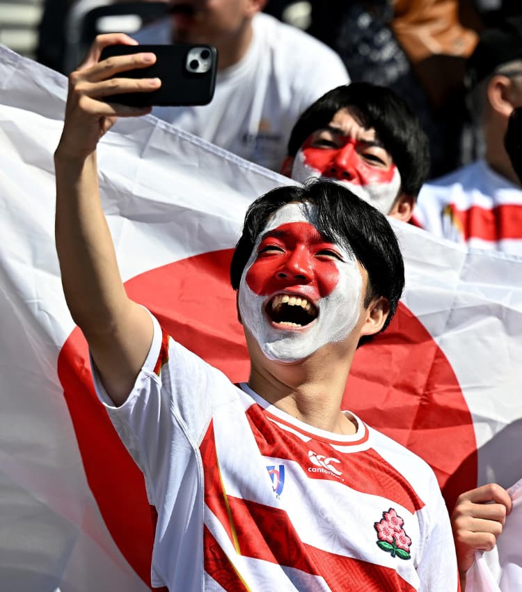 Japan rugby fans with Japanese flag face paint cheer and take a selfie in front of large Japanese flags in a stadium.