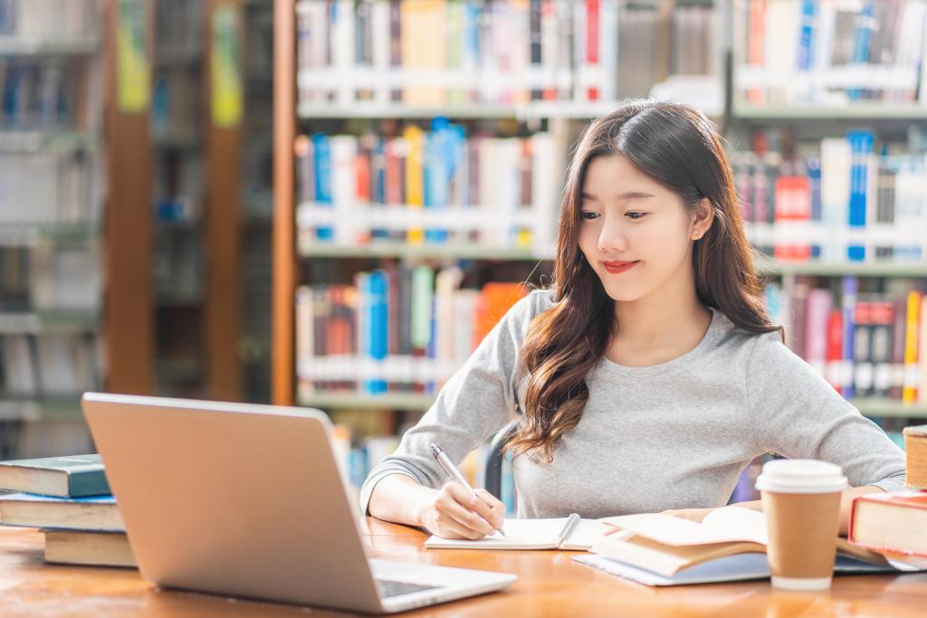 Girl in a library looking at a laptop and making notes